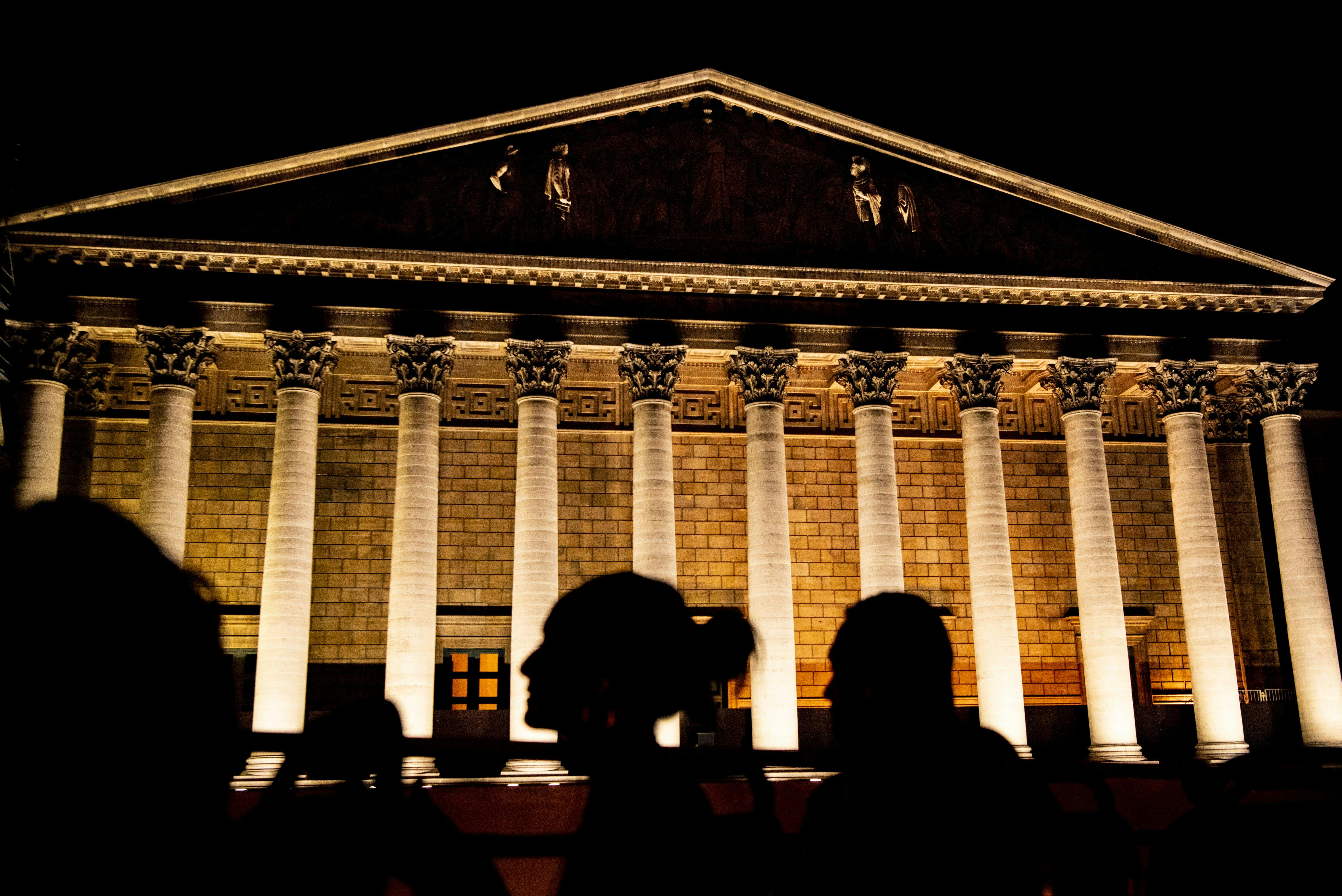 Osvětlená neoklasicistní budova se sloupy v noci, v popředí siluety dvou lidí.