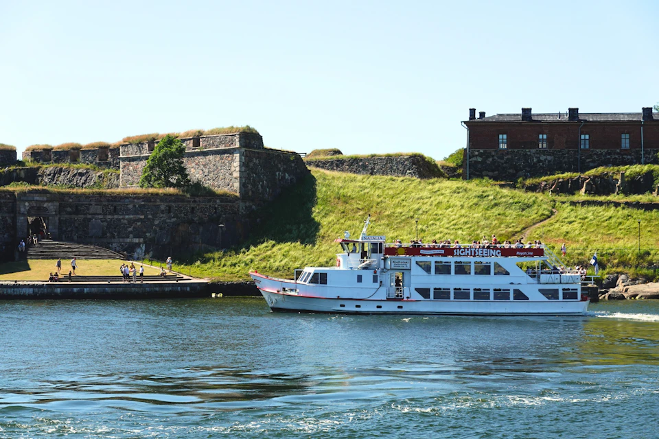 A white sightseeing boat sails near a grassy hillside with stone fortress walls and a brown building in the background.