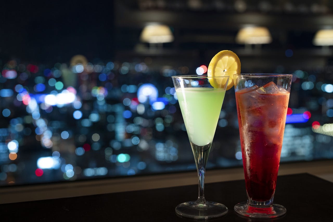 Два коктейля: зеленый напиток с ломтиком лимона и красный напиток с кубиками льда, с размытым городским пейзажем на заднем плане.