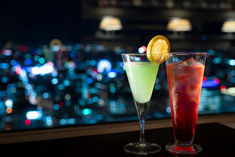 Two cocktails: a green drink with a lemon slice and a red drink with ice cubes, with a blurred cityscape in the background.