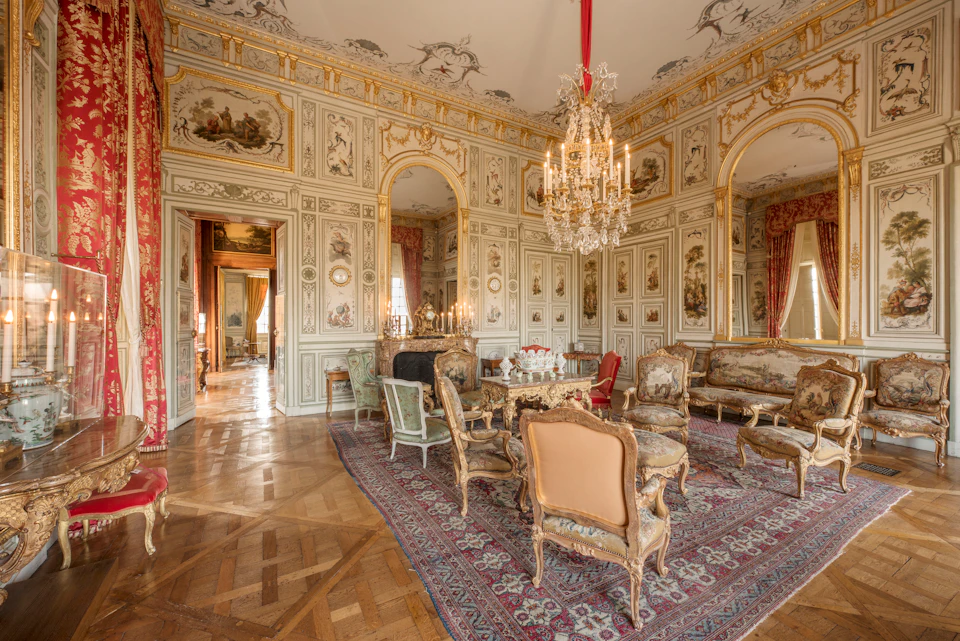 Ornate room with antique furniture, chandeliers, detailed wall panels, a fireplace, and large windows with red curtains.