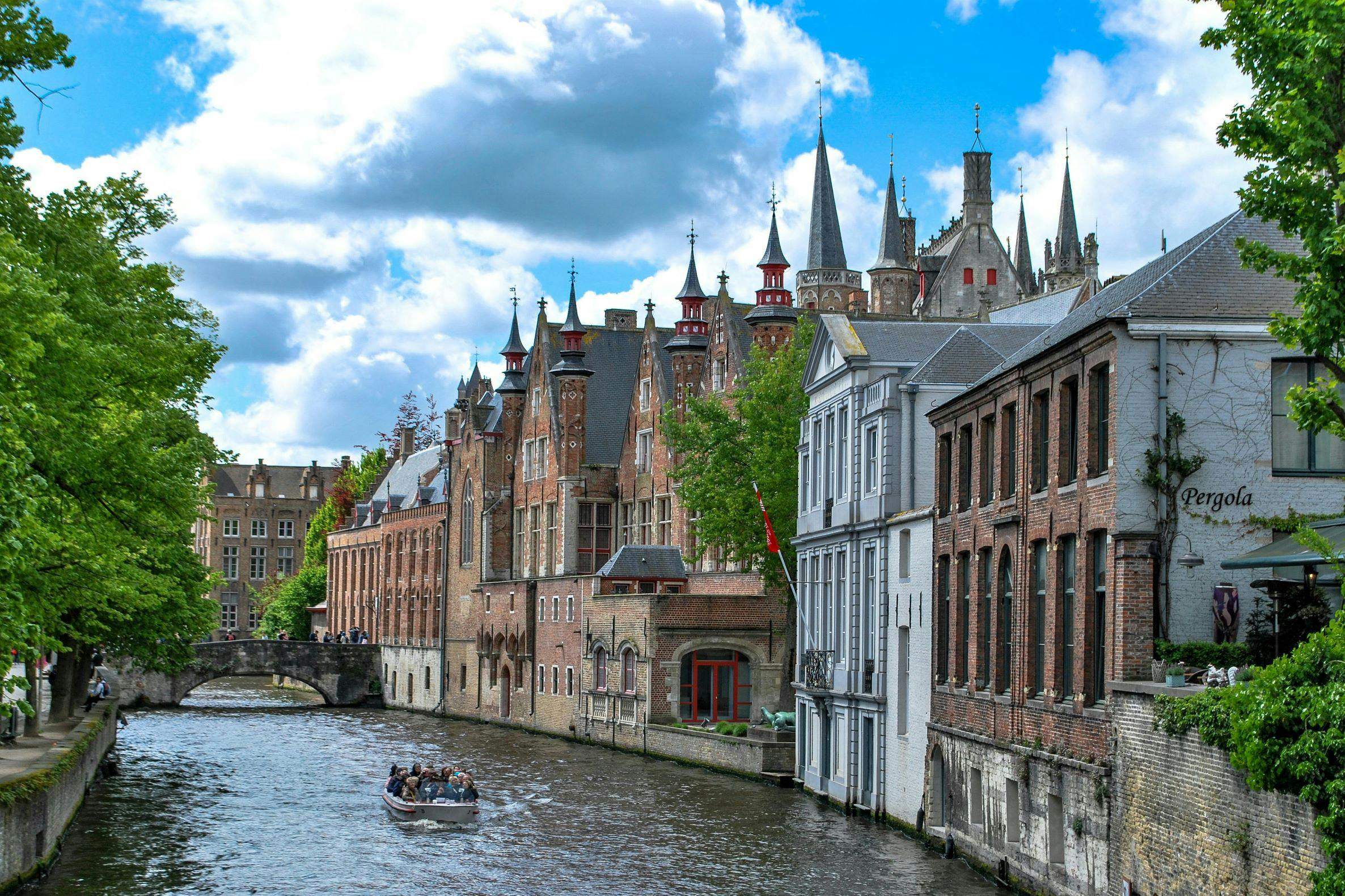 A canal with a boat carrying people, lined by historic buildings and a church with spires under a partly cloudy sky.