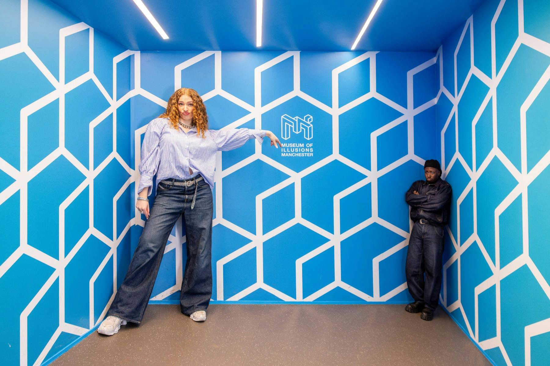 Två personer står i ett optiskt illusionsrum med blå geometriska väggar och texten "Museum of Illusions Manchester" synlig.