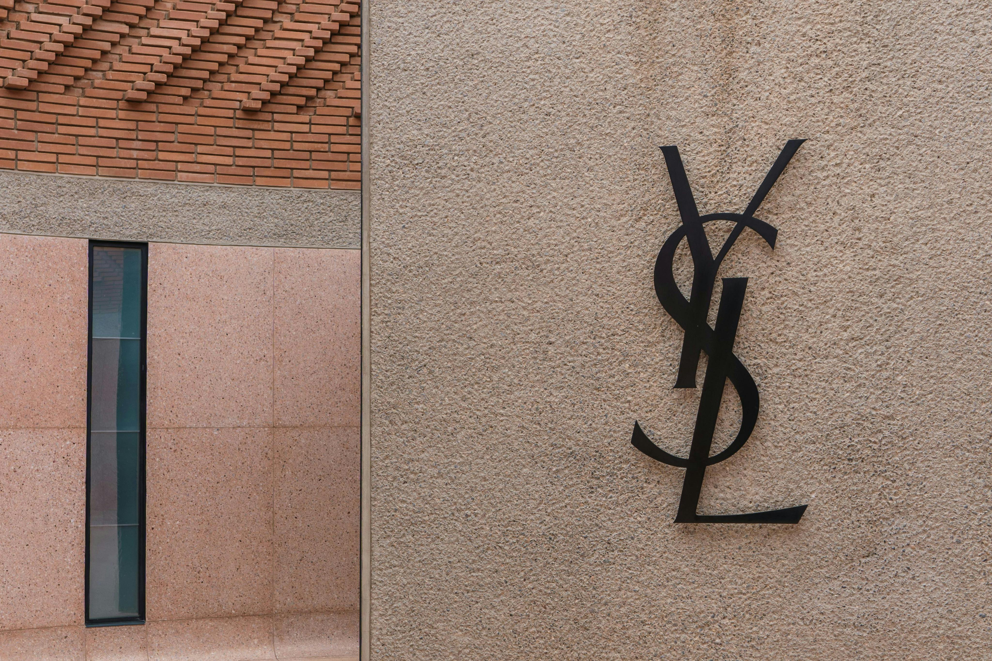 A wall with the Yves Saint Laurent logo in black on a beige textured surface, next to a tiled area and brick detail above.