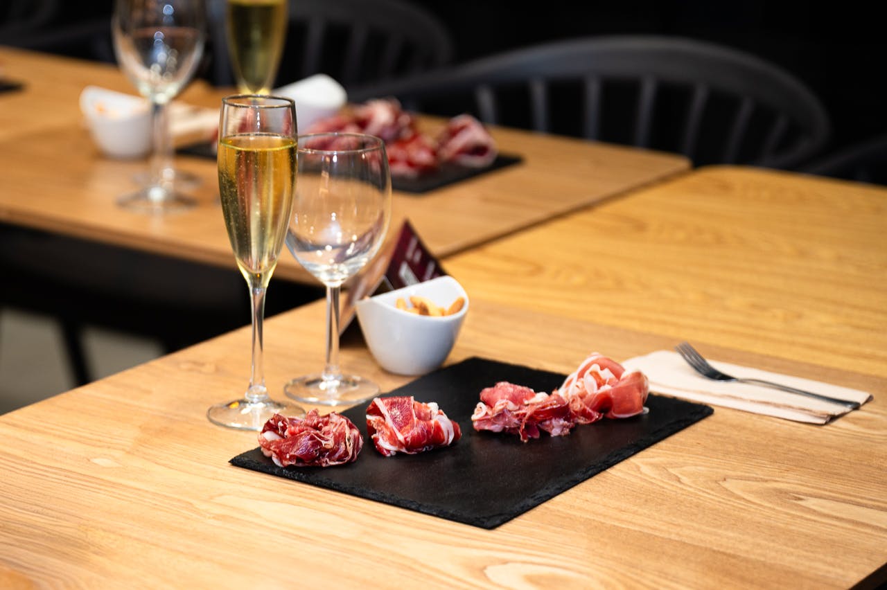 Et træbord med en skiferplade af hærdede kødskiver, et glas champagne, et tomt vinglas og en lille skål snacks.