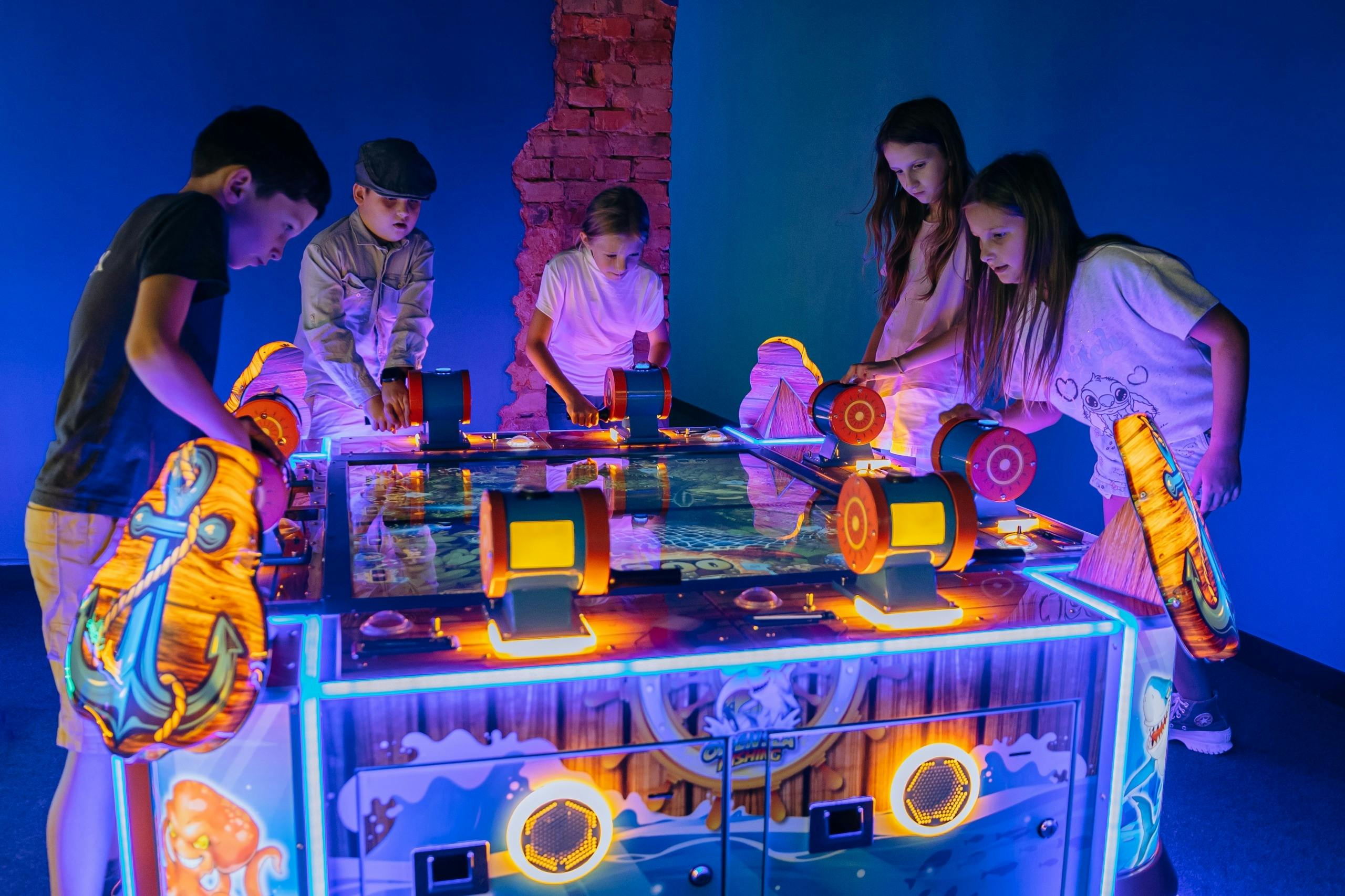 Three children play an air hockey game with illuminated paddles and a colorful table in a room with blue lighting.