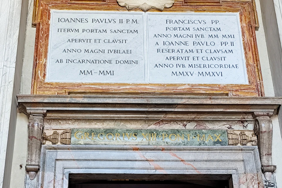 A marble plaque with Latin inscriptions above a doorway, including references to Pope John Paul II and Pope Francis.