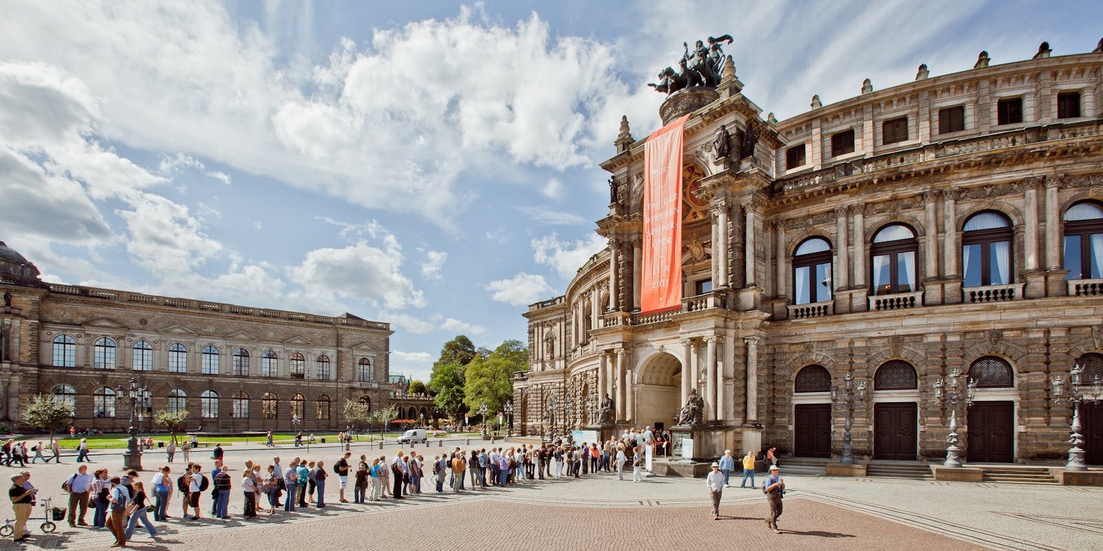 People line up outside an ornate historic building with a large orange banner and statues under a partly cloudy sky.