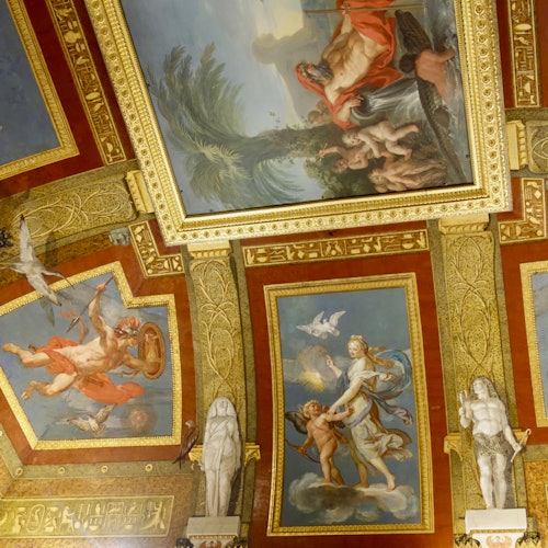 Soffitto ornato con dipinti dettagliati di scene mitologiche, circondato da decorazioni dorate e statue su uno sfondo rosso.