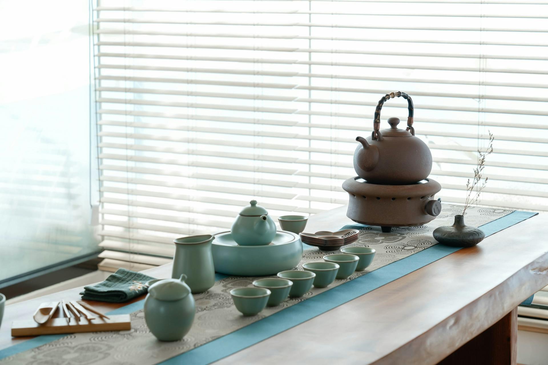 A tea set with cups and a teapot on a table, next to a kettle on a stove, with blinds in the background.