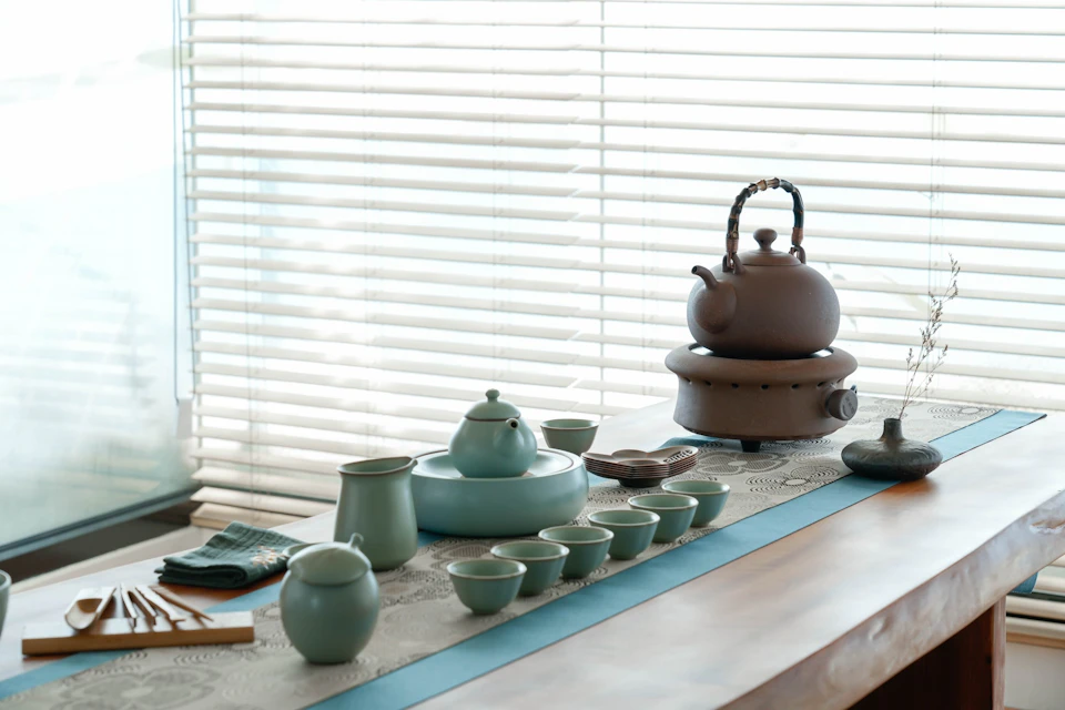 A tea set with cups, a teapot, and a kettle arranged on a table near a window with blinds.