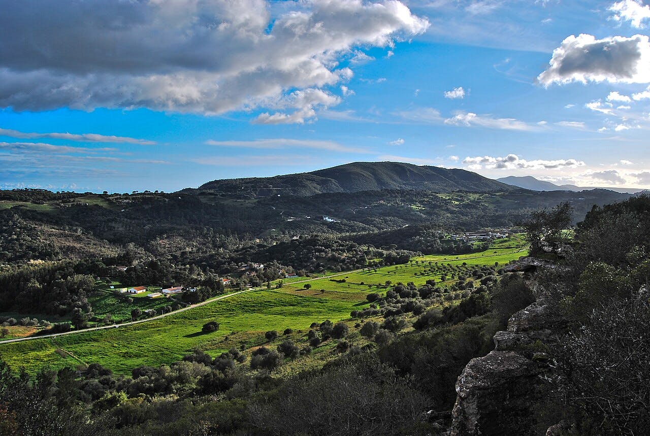 Paesaggio panoramico con campi verdi, alberi sparsi, piccoli edifici e colline ondulate sotto un cielo azzurro parzialmente nuvoloso.