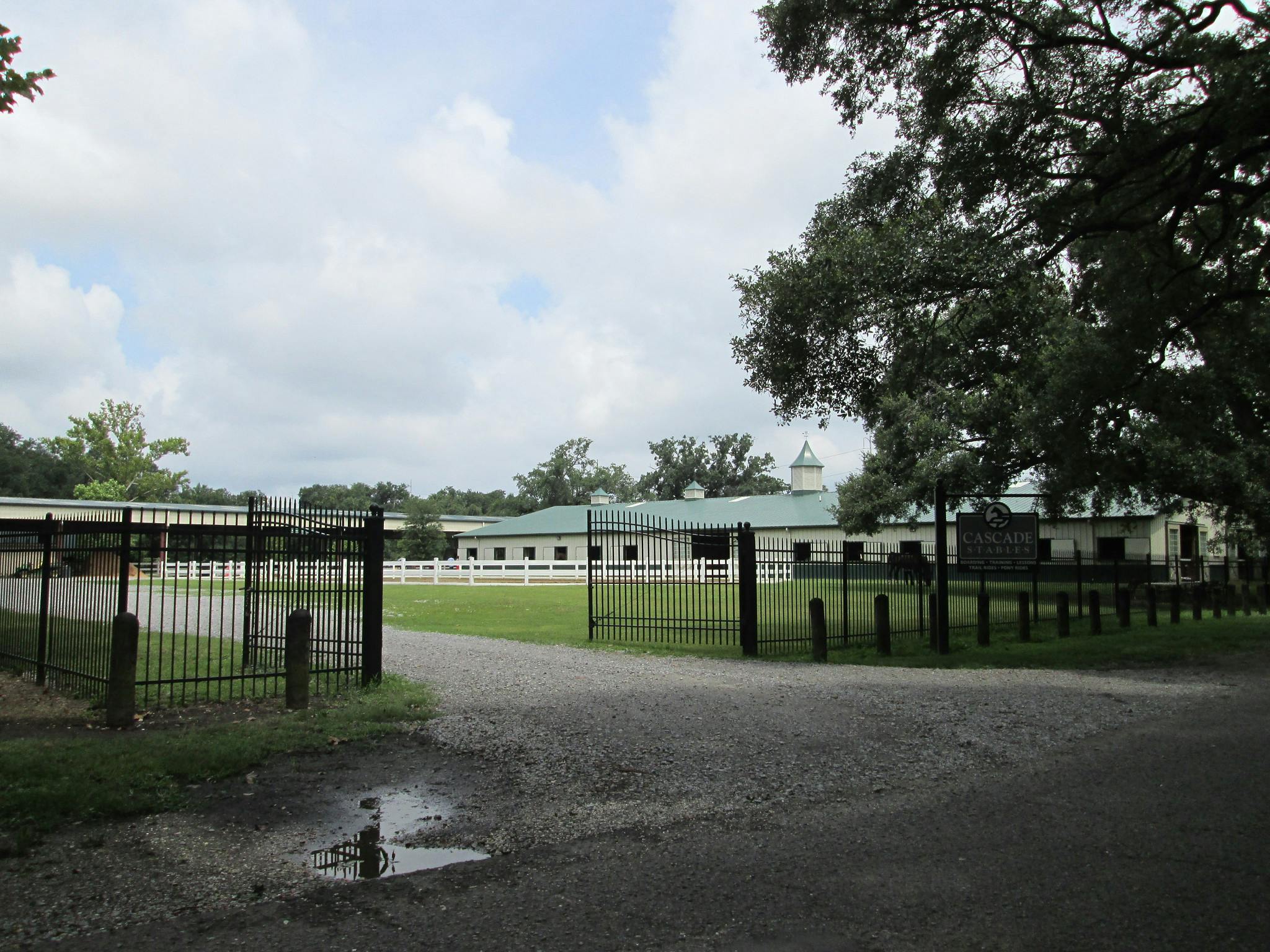 Indgang til Cascade Stables med en lukket indkørsel, sti med træer, hvidt hegn og bygning med grønt tag.