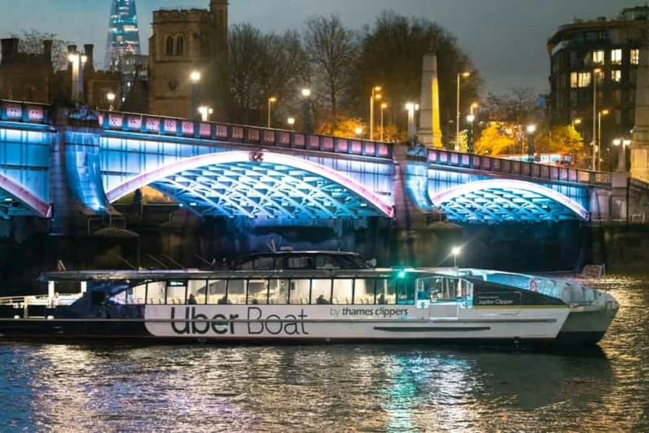 A boat labeled 'Uber Boat by Thames Clippers' on a river at night, near a lit-up bridge and trees with soft lighting.