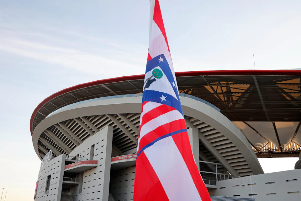 A red, white, and blue flag with a club emblem in front of a modern stadium structure under a clear sky.