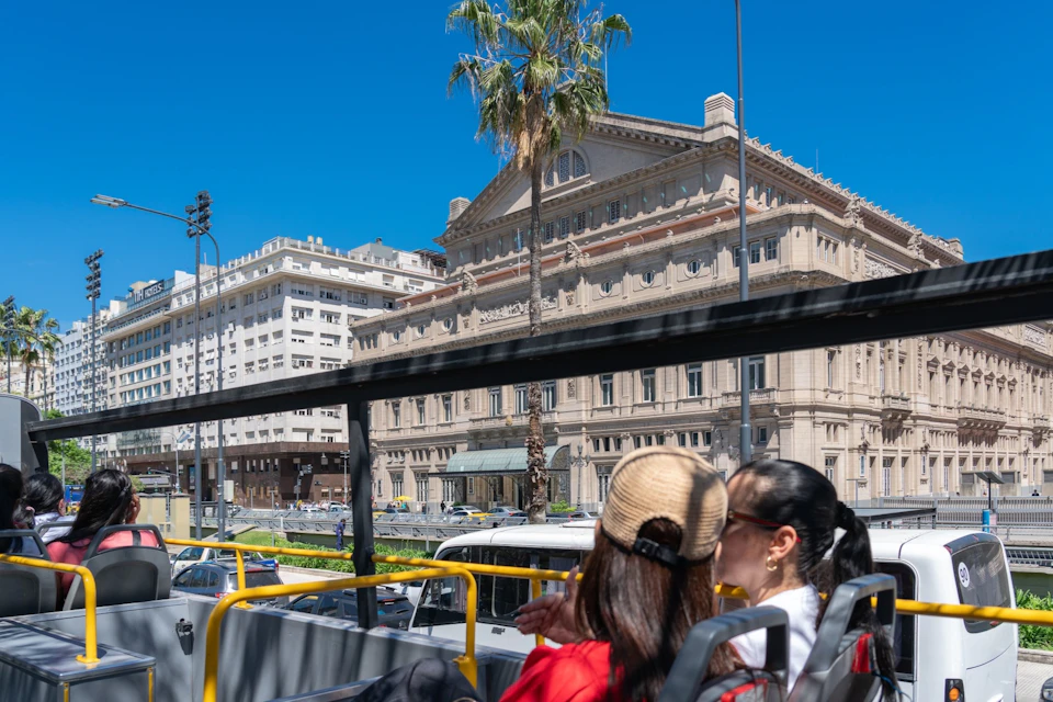 Two people on a tour bus view an ornate building and palm tree under a clear blue sky in a city setting.