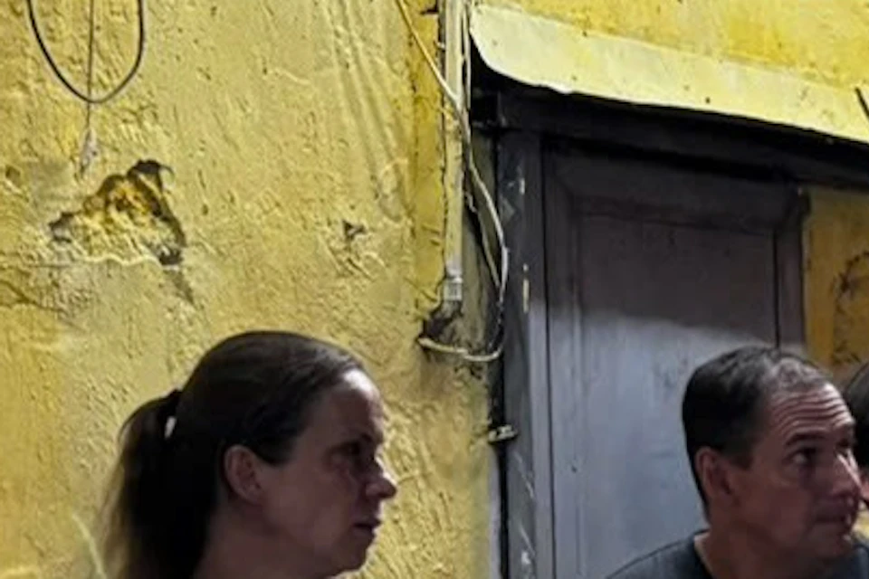 People sitting at small outdoor café tables against a yellow wall with hanging red flags, string lights, and overhead awning.