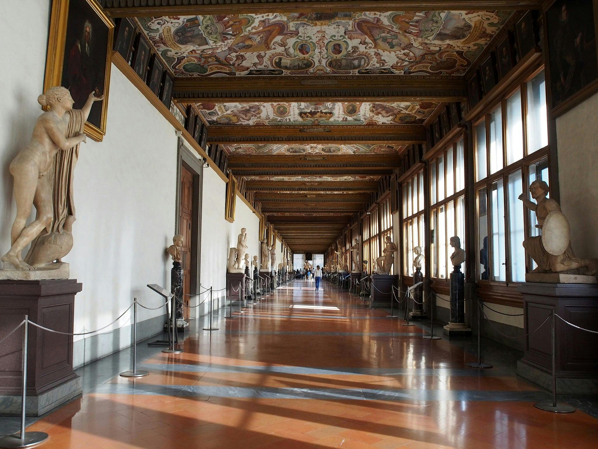 A long, ornate corridor with statues, paintings on the walls, and a decorated ceiling, lit by large windows on the right.