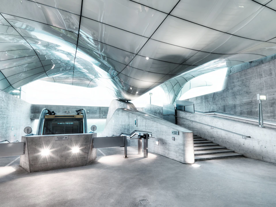 Modern, futuristic indoor space with sleek, reflective ceiling, concrete walls, no people, escalator, stairs, and turnstiles.