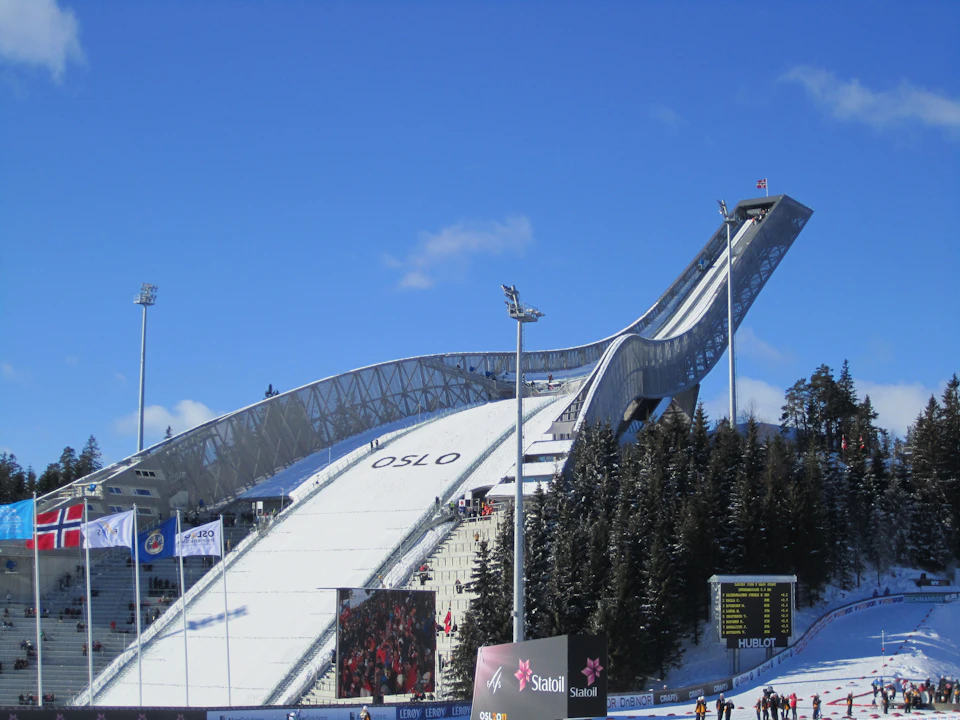 A large ski jumping hill labeled "OSLO," surrounded by snowy trees, spectators, and advertising boards, under a clear blue sky.