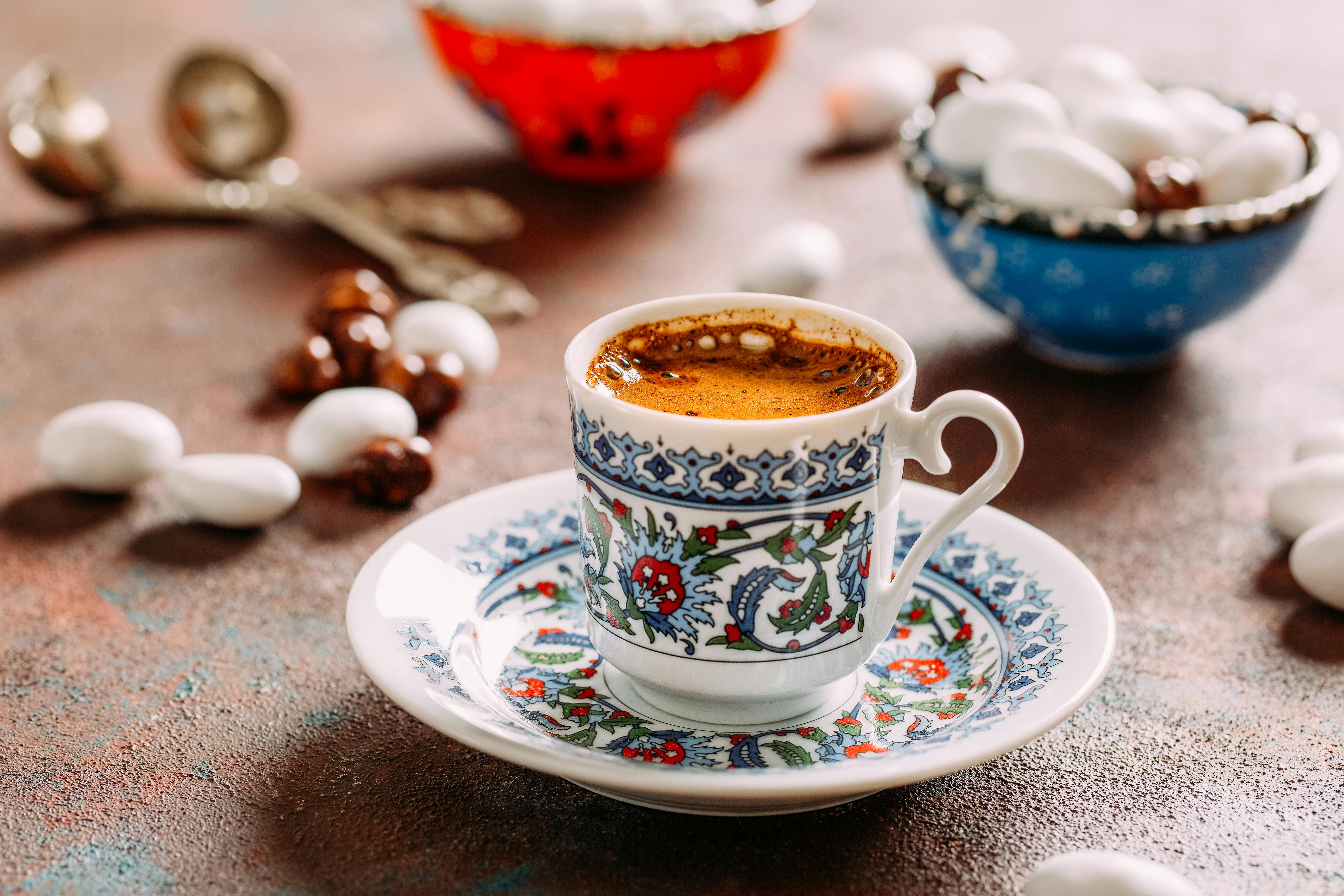 Decorative cup of espresso on a matching saucer, surrounded by nuts, white candies, and ornate bowls.