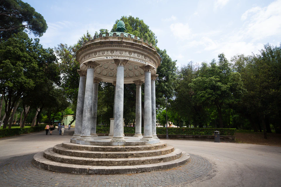 A circular stone pavilion with columns and a dome, surrounded by trees in a park setting under a cloudy sky.