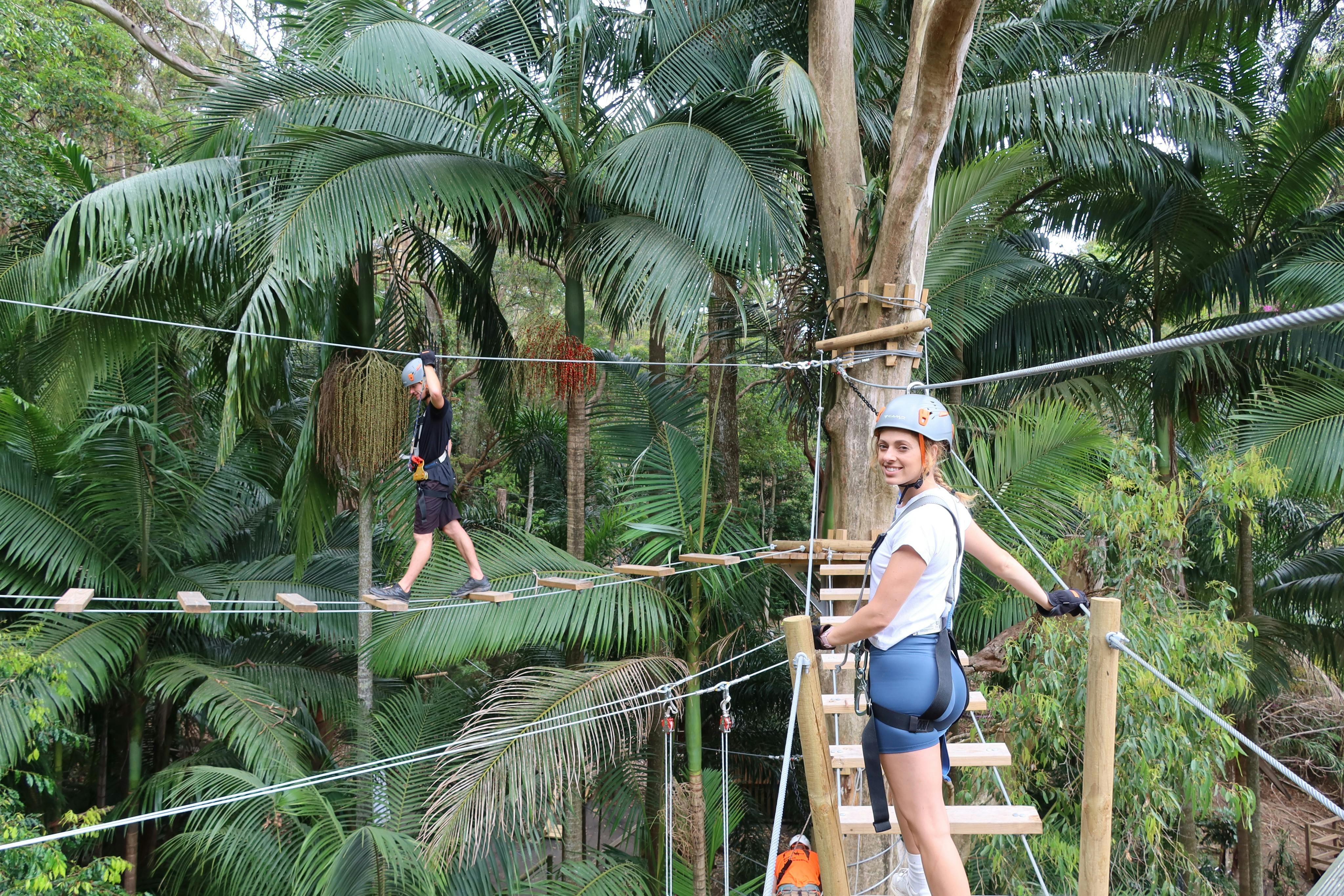 Ένα άτομο που φοράει κράνος και εξοπλισμό ασφαλείας στέκεται σε μια γέφυρα με σχοινιά σε ένα καταπράσινο, τροπικό δάσος.