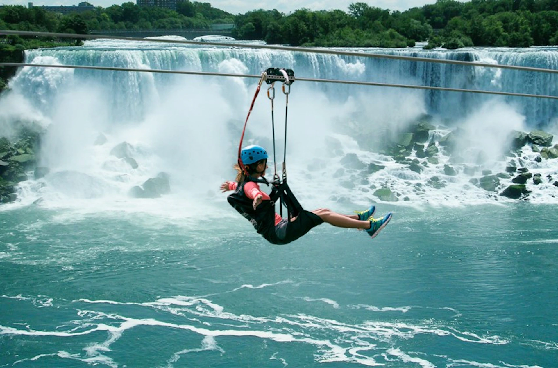 Person ziplining in front of a large waterfall, wearing a blue helmet and harness, with lush greenery in the background.