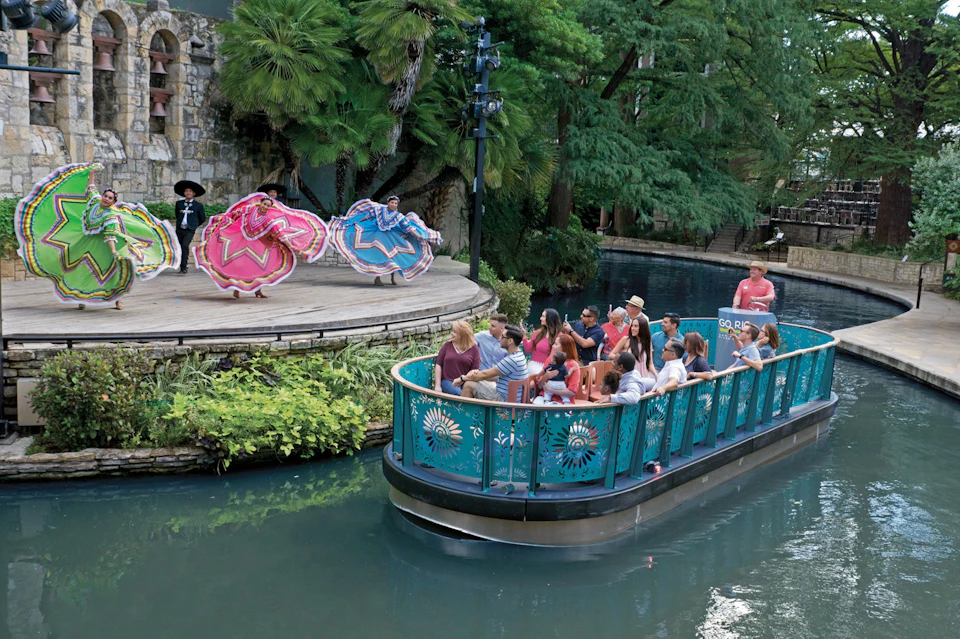 A group of people on a boat passing by a riverside stage with dancers in colorful dresses performing.