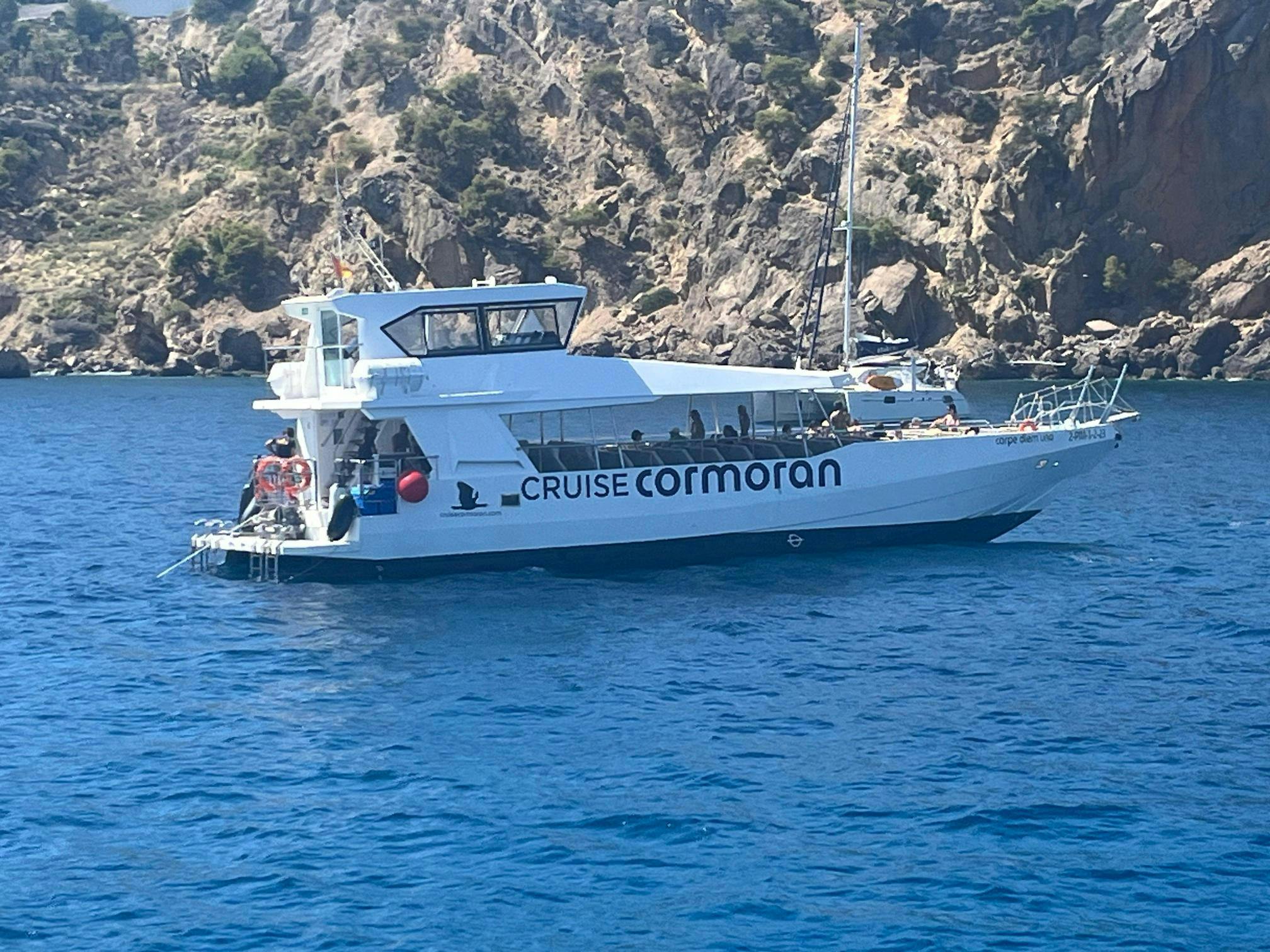 Una barca bianca con la scritta "Cruise Cormoran" galleggia sull'acqua blu vicino a scogliere rocciose con poca vegetazione.