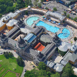 Flygfoto över ett stort spa-komplex med flera utomhuspooler, omgivet av genomarbetade historiska byggnader och grönska.