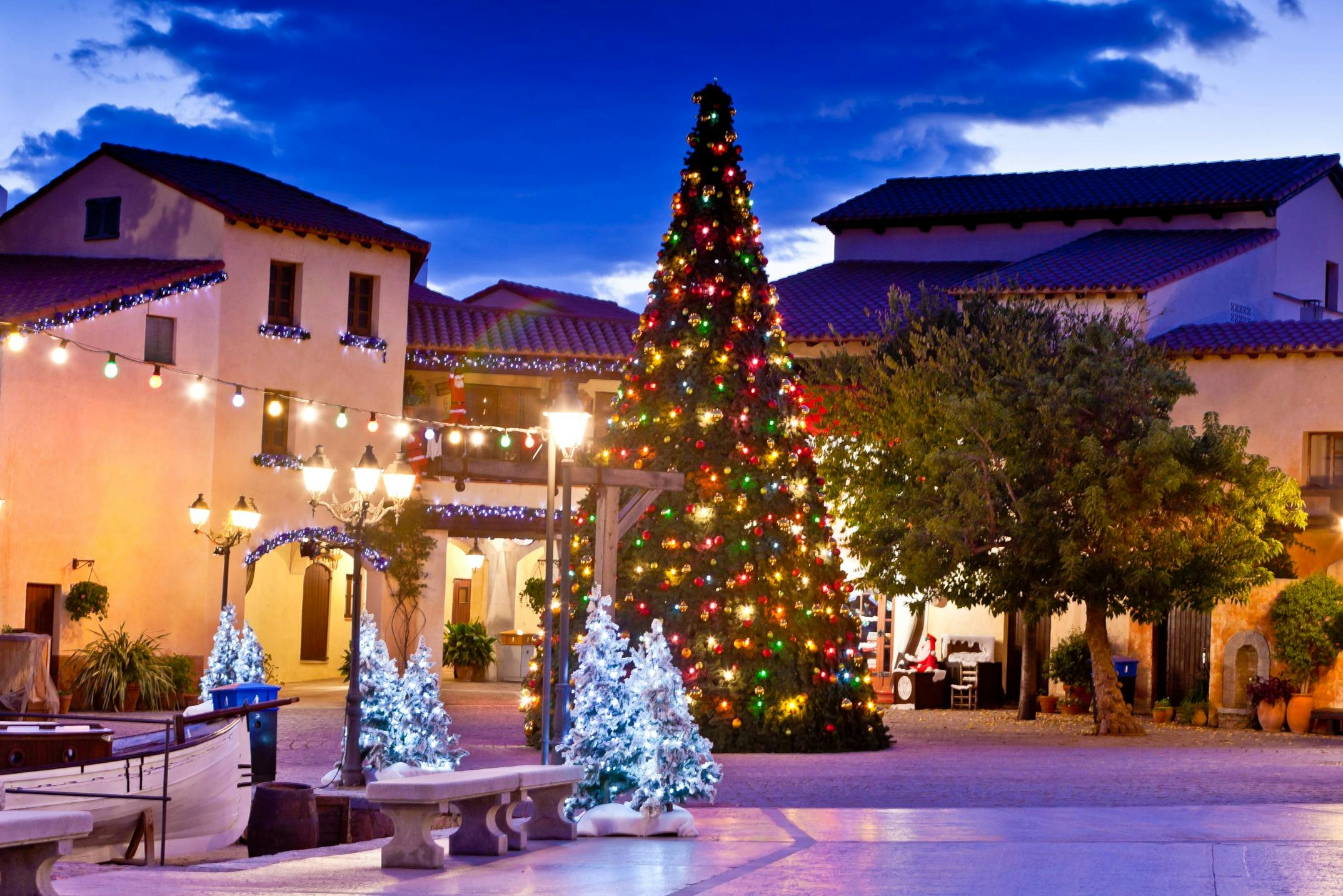 Cour décorée de manière festive avec un grand arbre de Noël, des arbres plus petits illuminés, des lanternes et des bâtiments illuminés sous un ciel crépusculaire.