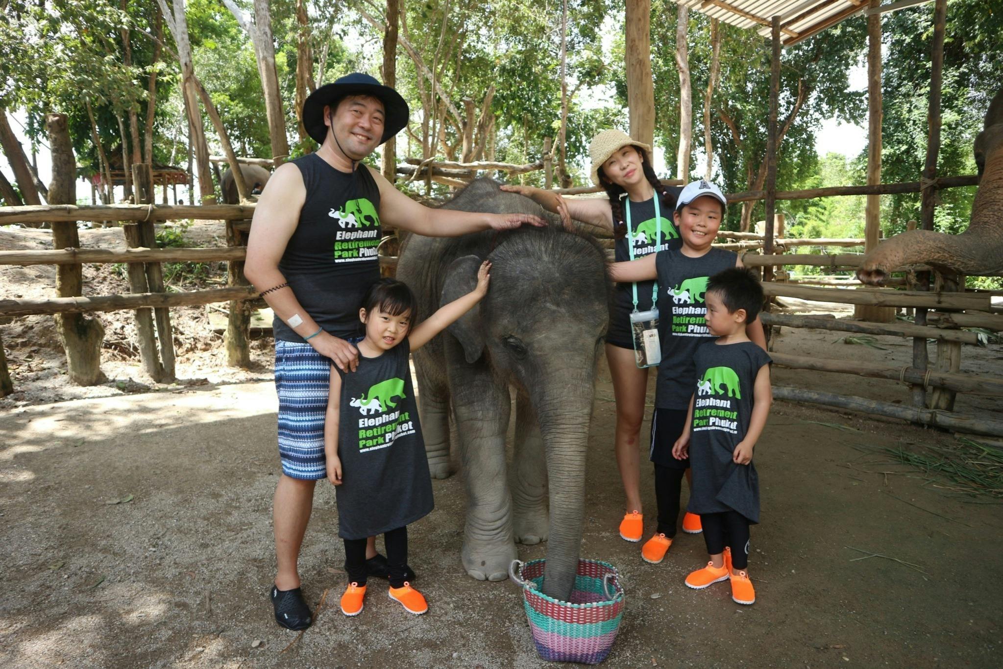 4명의 가족이 어울리는 셔츠와 주황색 신발을 신고 나무를 배경으로 인클로저 안에 코끼리 송아지를 쓰다듬고 있습니다.
