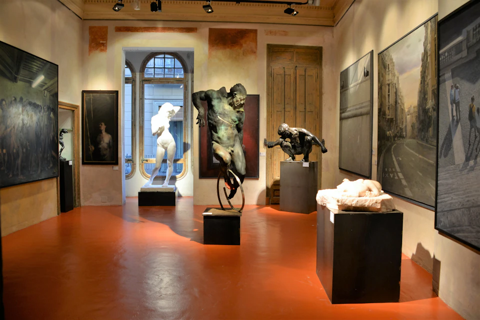 Gallery room with sculptures and paintings, including a prominent dark-colored statue of a muscular figure on a unicycle.