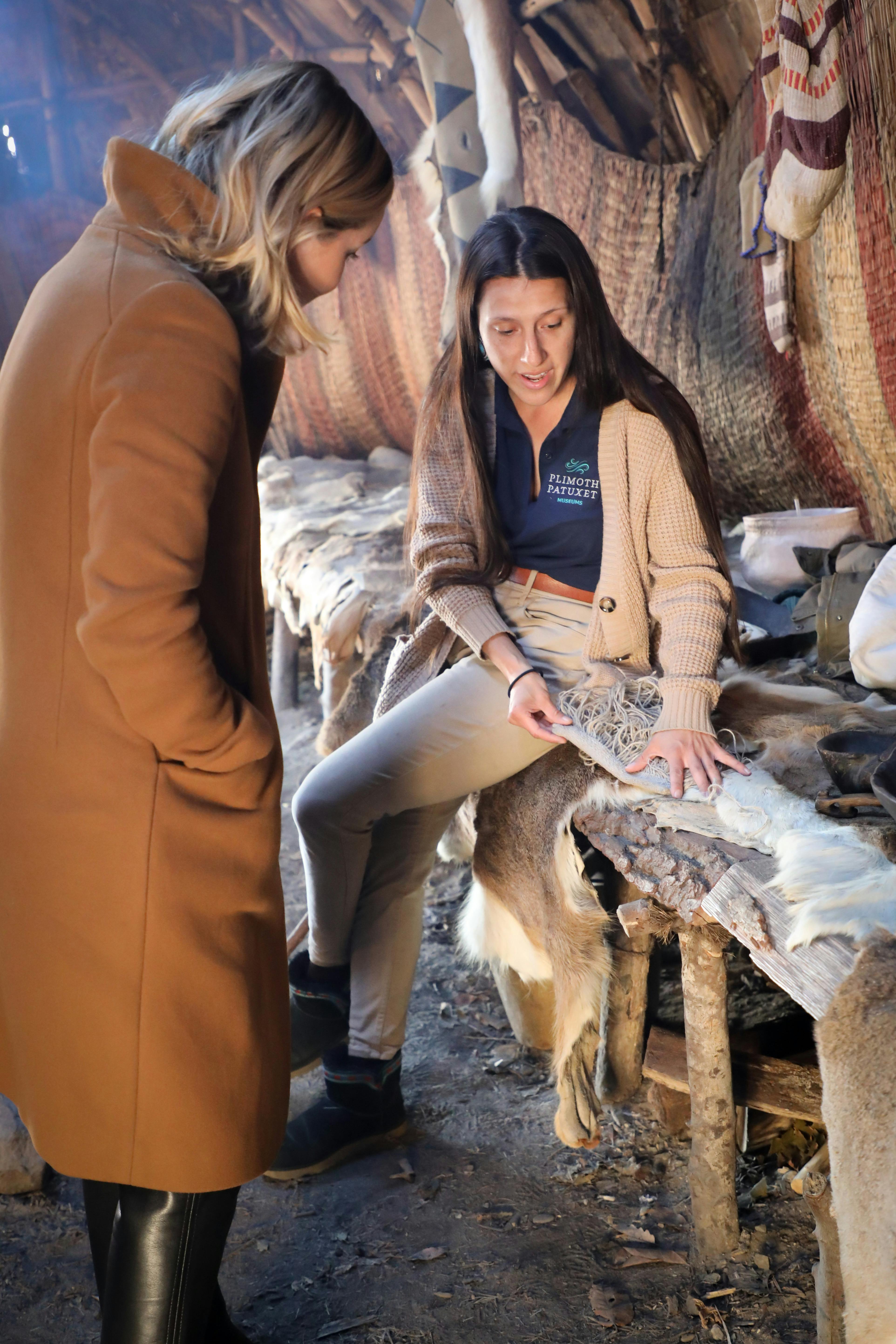 Dvě ženy v rustikálním útulku zkoumají textilie na dřevěné lavičce. Jedna žena ukazuje na látku a něco vysvětluje.