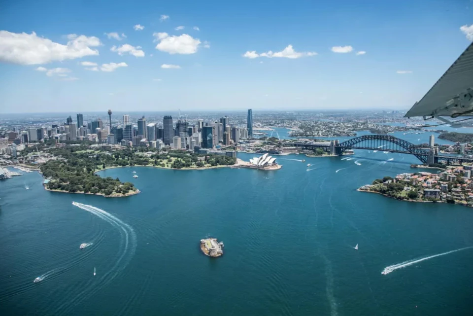 Aerial view of a city skyline by a harbor, featuring a prominent bridge and an iconic white architectural structure.