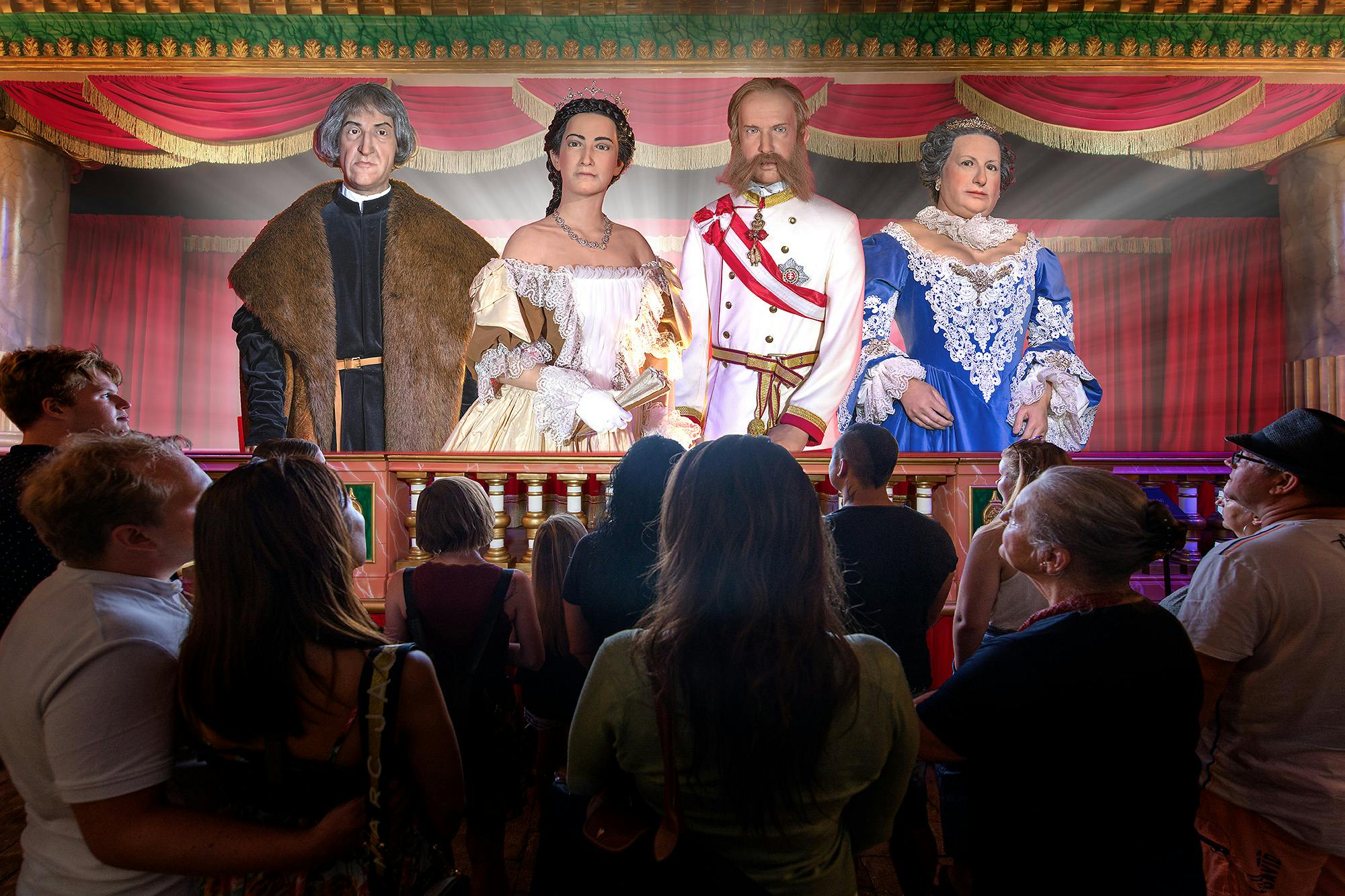 People observing life-like historical figures on a stage with ornate red curtains and golden decorations.