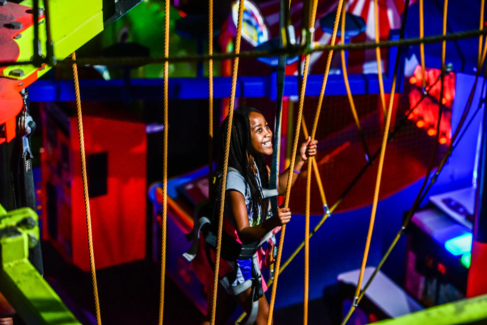 A girl smiling while navigating a ropes obstacle course indoors, surrounded by bright and colorful elements.