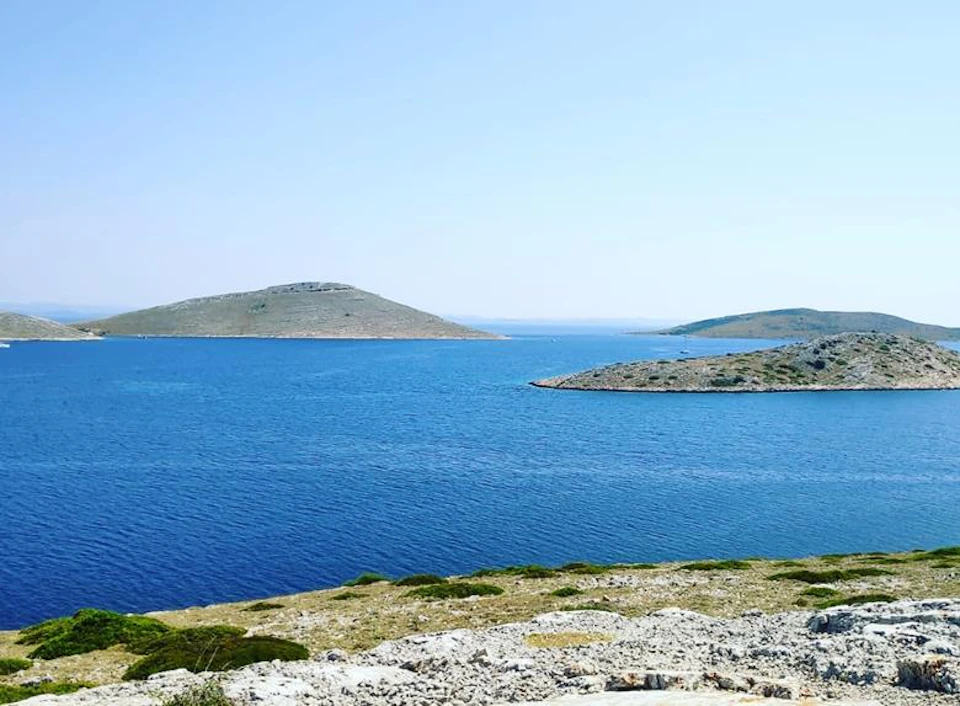 Serene landscape with calm blue sea, sparse vegetation, and rocky hills under a clear, blue sky.