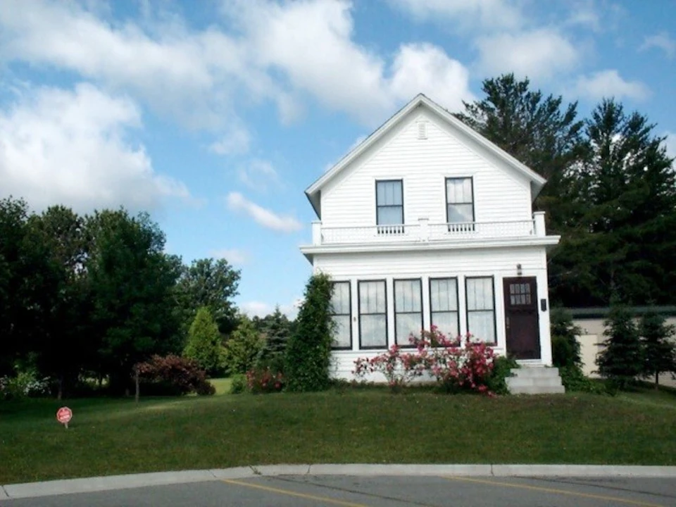 Two-story white house with a porch, surrounded by green lawn, bushes, and trees, under a partly cloudy sky.