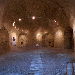 Ett svagt upplyst stenväggigt rum med välvda dörröppningar, en konstutställning och en central cirkulär ljusarmatur.