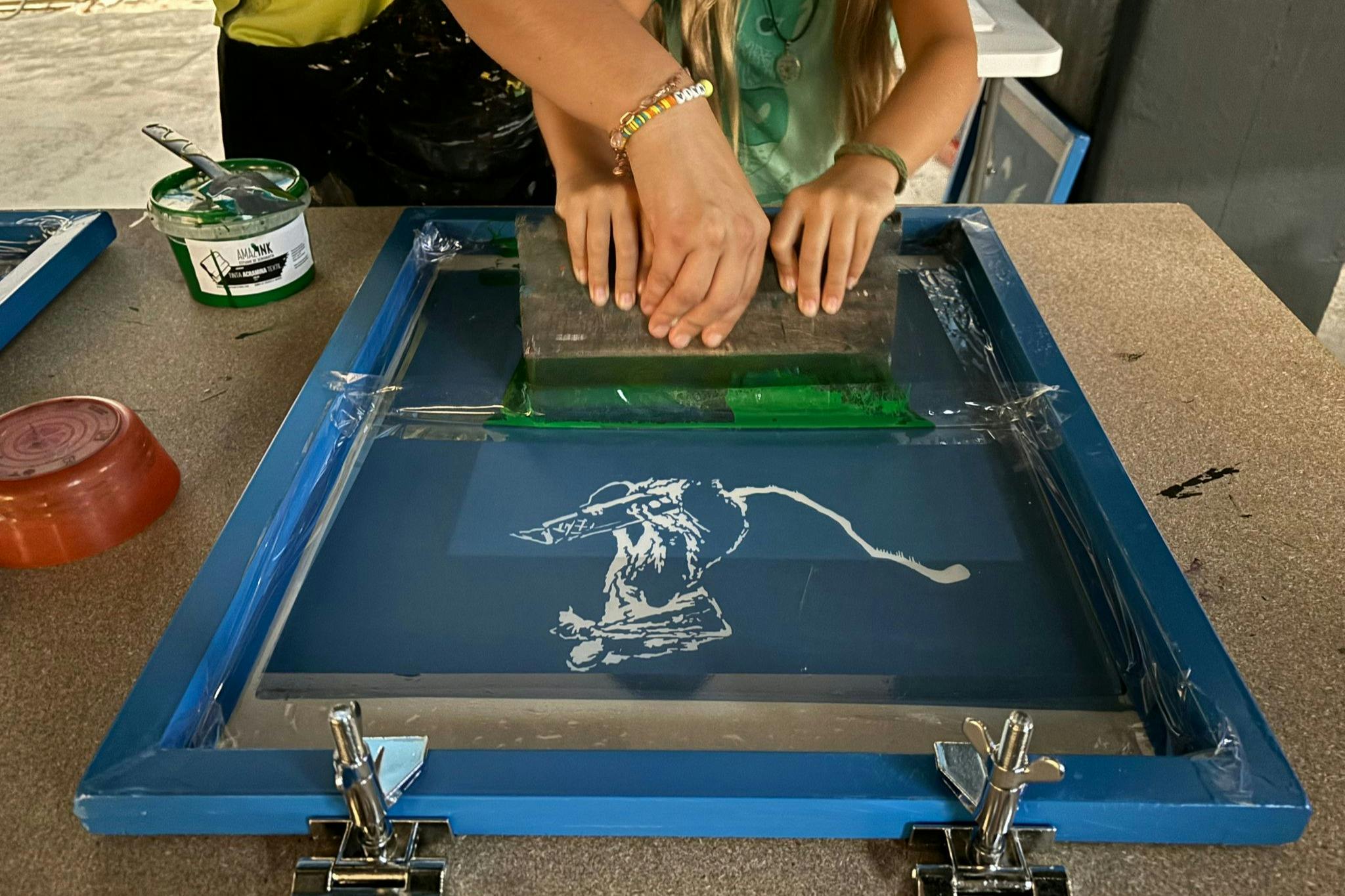 Dues persones utilitzant un marc de serigrafia amb una rasqueta per aplicar tinta verda a un disseny d'una figura a la pantalla.