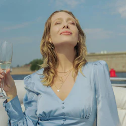A woman in a light blue dress sitting outdoors, holding a glass, with her face tilted upwards towards the sun.