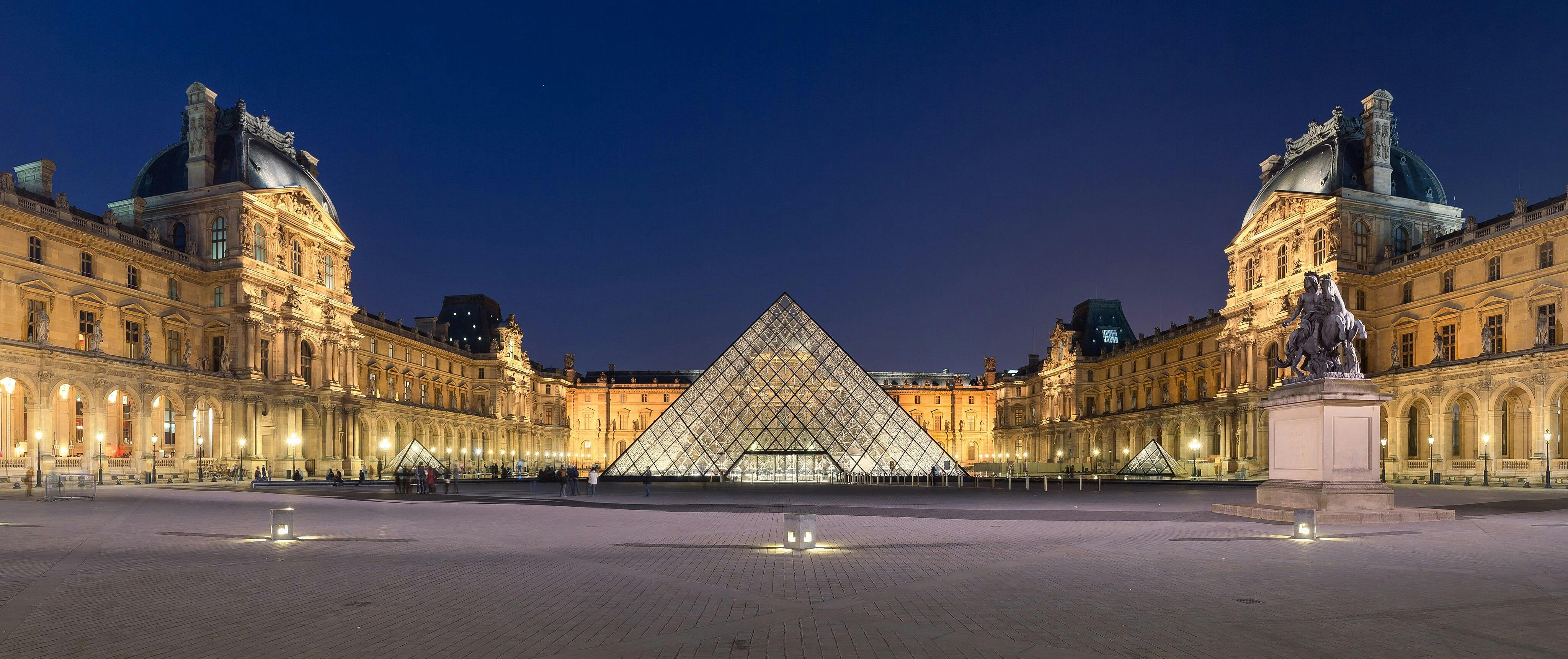 Beleuchteter Innenhof des Louvre-Museums mit zentraler Glaspyramide, historischen Gebäuden auf beiden Seiten und einem Reiterstandbild bei Nacht.