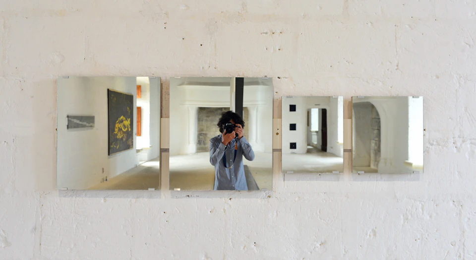 Person taking a photo of themselves in three mirrors mounted on a white wall in an art gallery.