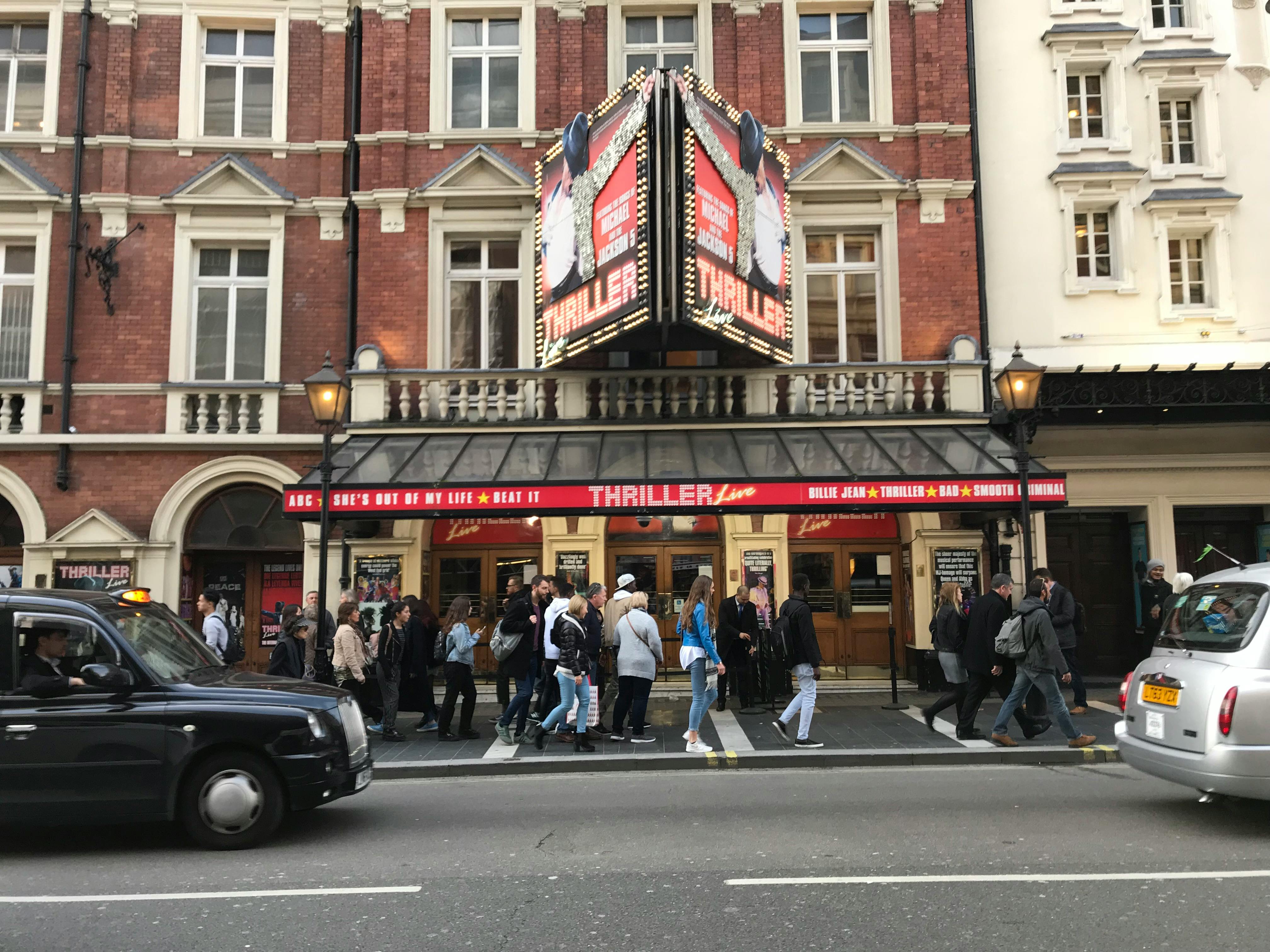 Folk samlades utanför en teater med ett tält som annonserade "Thriller Live", som ligger på en livlig gata med förbipasserande bilar.