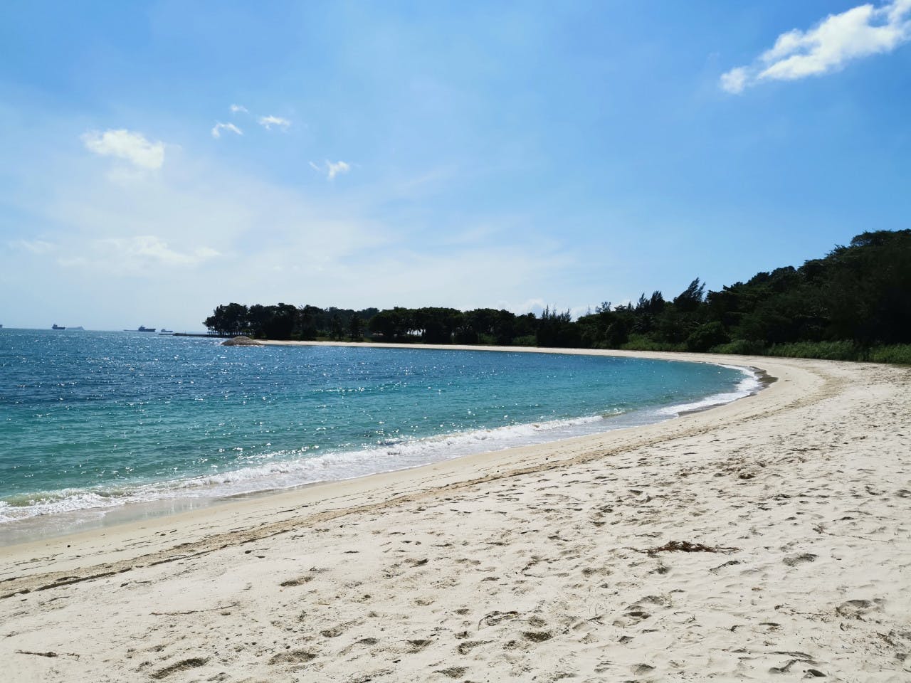 Klidná pláž s lehkým pískem, klidnou tyrkysovou vodou a vzdálenými stromy pod jasně modrou oblohou.