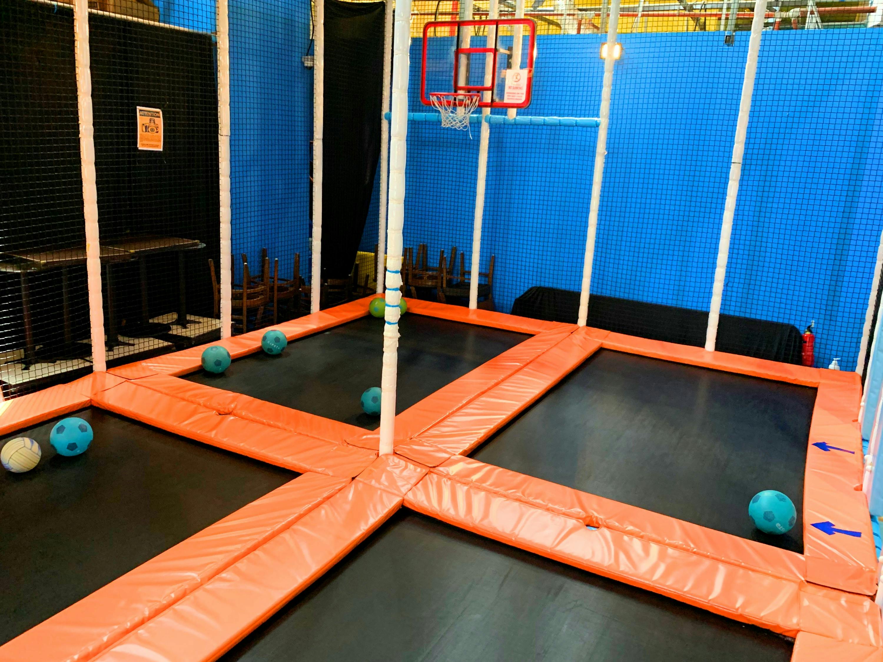 Indoor trampoline area with orange borders, blue walls, and a basketball hoop. Several soft balls are on the trampolines.
