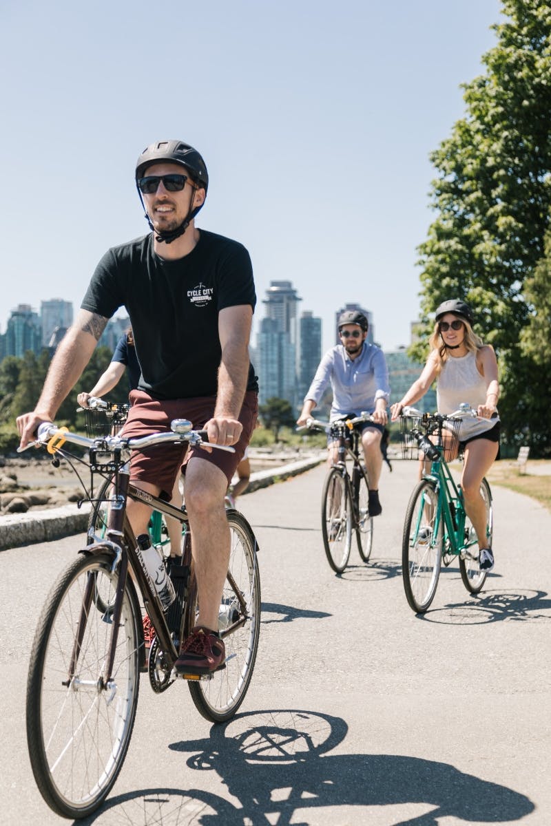 街のスカイラインと大きな木々を背景に、舗装された道を3人で自転車に乗っています。