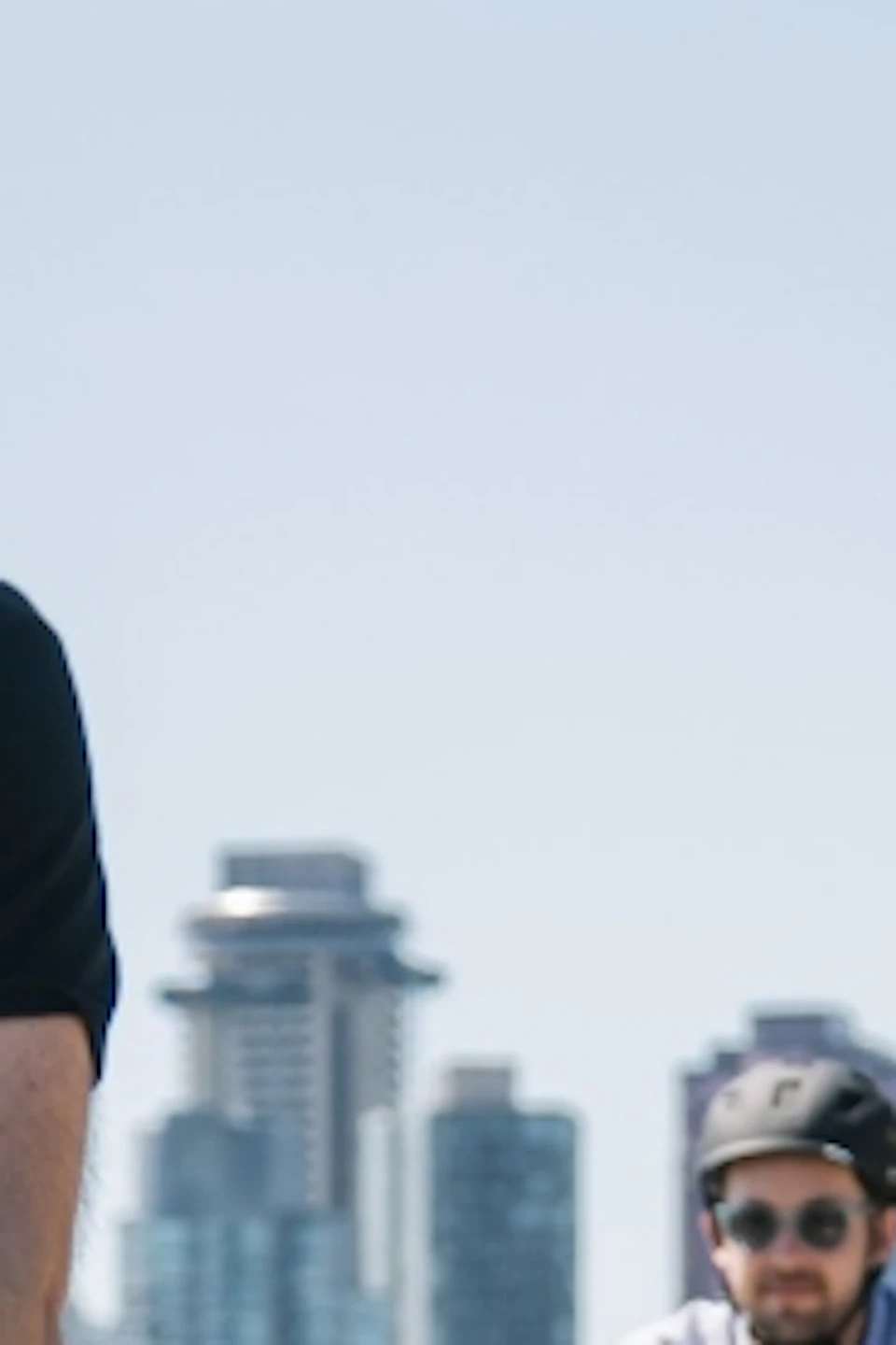 Three people ride bicycles on a paved path with a city skyline and large trees in the background.
